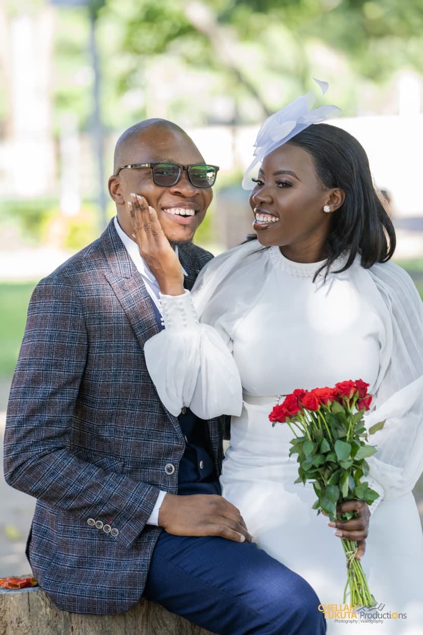 Playful couple moment - bride touching groom's face, both laughing with red roses